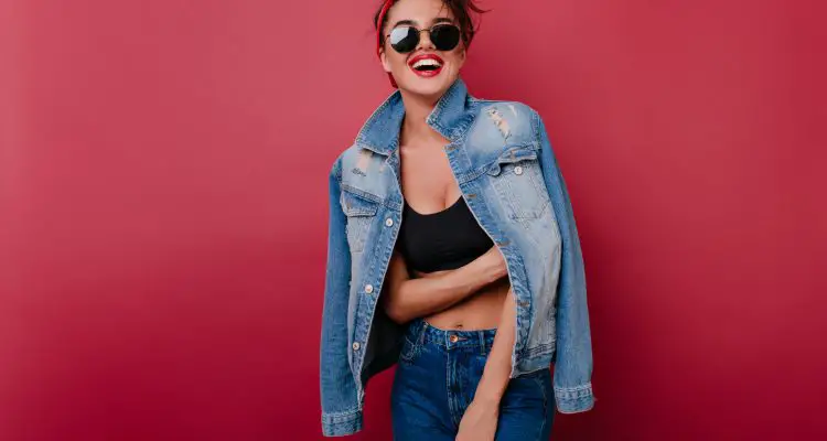 A women wearing a denim jacket, denim jeans, black crop top with headband and retro sunglasses laughing