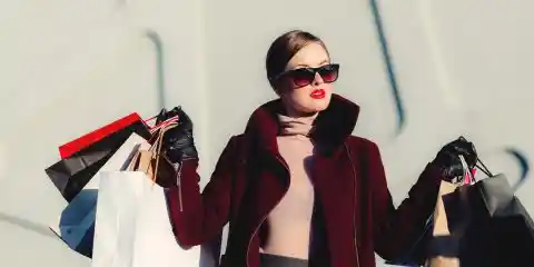 Women wearing pink turtleneck with retro sungalsses, grey skirt and maroon coat with lots of shopping bags in her hands