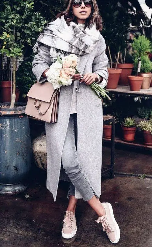 Women wearing long light grey winter coat with faded grey jeans, sneakers and chunky scarf with light pink handbag. 